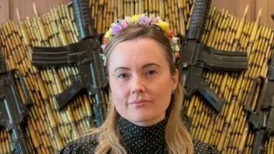 Uma mulher branca de cabelos loiros, a deputada Julia Zanatt (PL), usa uma tiara de flores coloridas e uma blusa preta de gola alta com bolinhas brancas e um colar de cruz prateada. Ela está posicionada à frente de um painel decorativo composto por dezenas de cartuchos de munição dispostos em arco e quatro fuzis pretos dispostos nas diagonais.